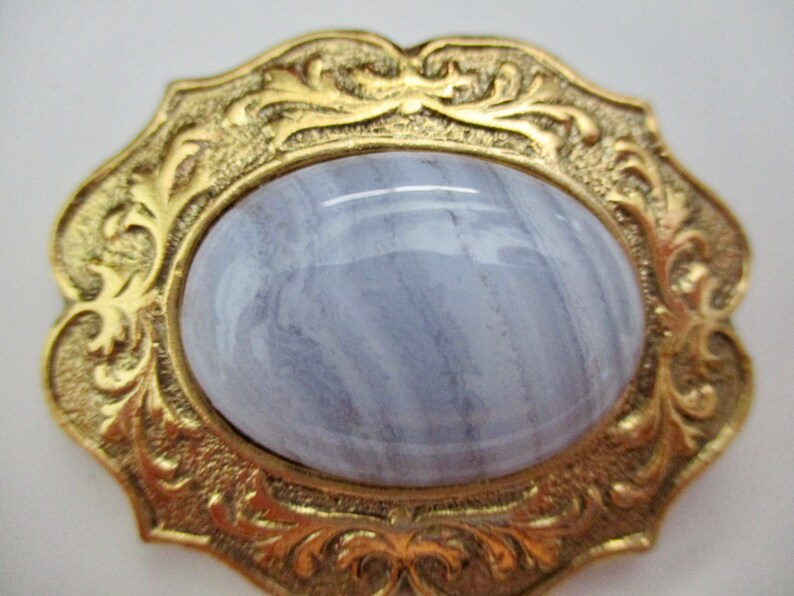 May include: A vintage gold-toned brooch with an oval, pale blue gemstone center. The gemstone has a marbled appearance. The brooch has an ornate, floral design around the gemstone.