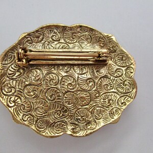 May include: A vintage gold-toned brooch with an oval shape and a decorative, swirling pattern. The back of the brooch features a pin clasp for secure attachment. The brooch is a classic accessory.