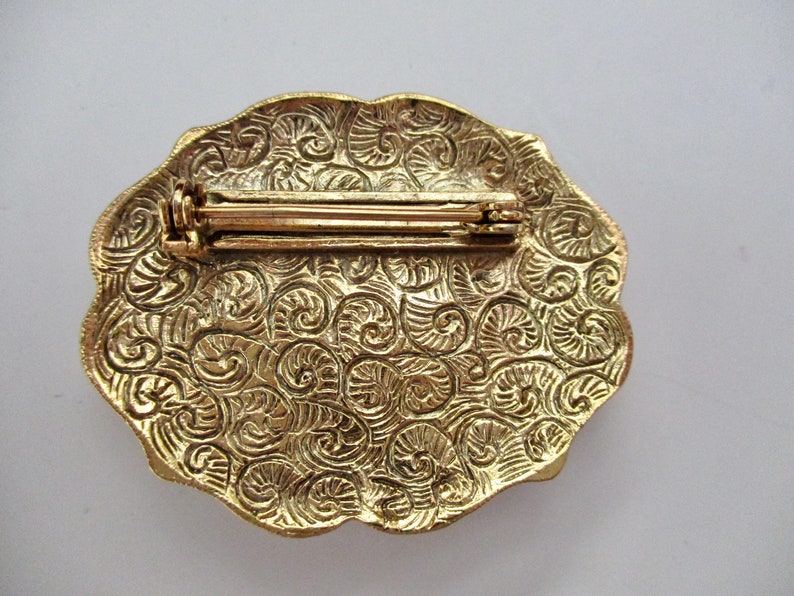 May include: A vintage gold-toned brooch with an oval shape and a decorative, swirling pattern. The back of the brooch features a pin clasp for secure attachment. The brooch is a classic accessory.