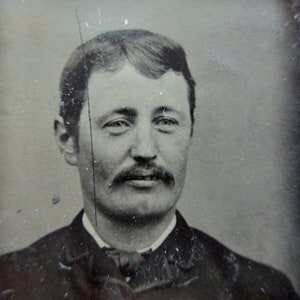 May include: A vintage black and white portrait of a man with a mustache, wearing a dark suit and a collared shirt. The image is slightly damaged, with scratches and imperfections, giving it an antique appearance. The man has short hair and a serious expression.