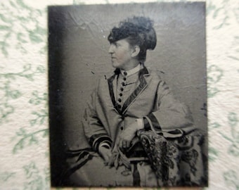 May include: A vintage black and white photograph of a person in profile, wearing a dark hat and a coat with dark trim. The coat has a high collar and button details. The person is seated, with a patterned fabric visible.