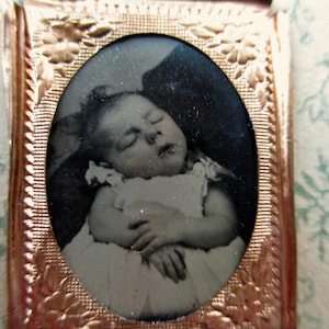 May include: Antique black and white photograph of a sleeping baby in a white dress, set within an oval frame. The frame is a detailed, ornate gold color, with floral patterns. The background is a light blue floral pattern.