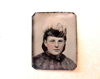 May include: Antique portrait of a person wearing a hat with feathers and a fur-trimmed coat. The image is a vintage photograph with a faded appearance, showing the subject's face and upper body. The photograph has a rectangular shape with rounded corners.