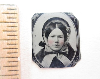 May include: A vintage tintype photograph of a person wearing a bonnet and a bow, displayed next to a wooden ruler showing a height of approximately 2 inches. The portrait is oval-shaped with a decorative black border and features hand-tinted pink cheeks.
