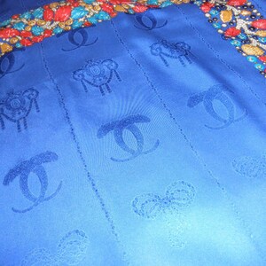 May include: A blue silk scarf with a repeating pattern of the Chanel logo, bows, and ornate designs. The scarf has a border of red, gold, and blue beads.