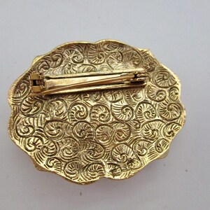 May include: Gold-toned vintage brooch with an oval shape and a decorative, swirling pattern. The brooch has a pin closure on the back. The edges of the brooch are scalloped.