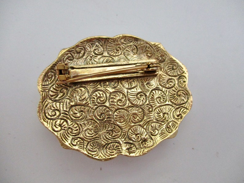 May include: Gold-toned vintage brooch with an oval shape and a decorative, swirling pattern. The brooch has a pin closure on the back. The edges of the brooch are scalloped.