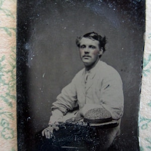 May include: A vintage black and white portrait of a person seated, wearing a light-colored jacket and a hat. The person is looking directly at the camera. The image has a grainy texture and is framed by a dark border.