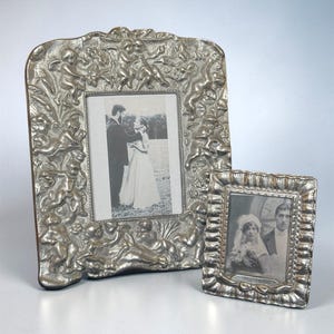 May include: Two silver-colored picture frames. The larger frame has an ornate design with cherubs, and the smaller frame has a decorative bow at the bottom. Both frames hold black and white photographs of couples.