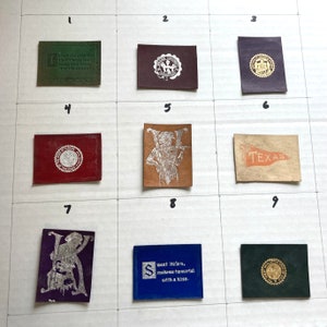 May include: Nine small, rectangular leather patches with various designs and text. The patches are arranged in a grid of three rows and three columns. The patches feature a variety of designs, including a seal, a flag, a musical instrument, and a letter. The text on the patches includes "slept and dream", "TEXAS", "sweet Helen, make me immortal with a kiss", and other phrases.