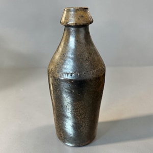 May include: A dark brown, antique-style bottle with a textured surface. The bottle has a narrow neck and a wider base, with a slightly flared lip. The bottle's color is a mix of dark brown and bronze.