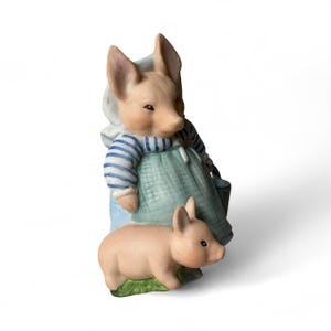May include: A ceramic figurine of a pig wearing a blue and white striped shirt and a green apron, holding a small bucket. A smaller piglet stands in front of the larger pig on a patch of green grass. The figurine has a light pink color.
