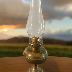 May include: A brass oil lamp with a clear glass chimney. The lamp is sitting on a wooden table.