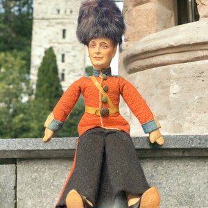 May include: A vintage doll dressed in a red and black uniform with a black fur hat. The doll is sitting on a stone ledge.