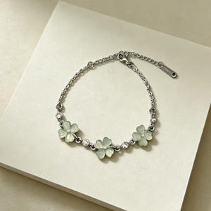 Puede incluir: Pulsera plateada con cuatro colgantes de flores verde pálido y detalles de cristal transparente. La pulsera está conectada por una fina cadena y se apoya sobre una superficie neutra.