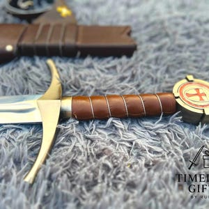 May include: A decorative sword with a brown leather-wrapped handle and a gold-colored crossguard. The pommel features a red cross design. The blade is silver and the scabbard is visible in the background. The sword is a Timeless Gift.