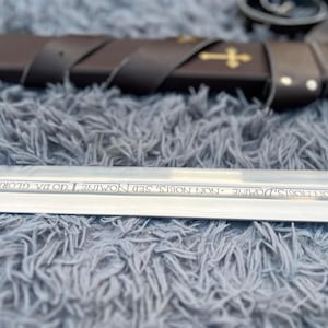 May include: A close-up of a sword with a silver blade and a brown leather sheath. The blade has an inscription. The sheath is decorated with gold crosses. The sword is resting on a gray, fuzzy surface.