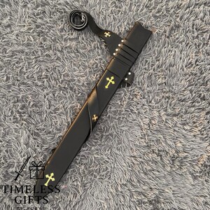 May include: A black sword with a leather sheath, featuring gold cross accents. The sword has a curved handle and a strap. The sheath is adorned with gold crosses. The text "TIMELESS GIFTS BY RUBAH" is visible at the bottom.