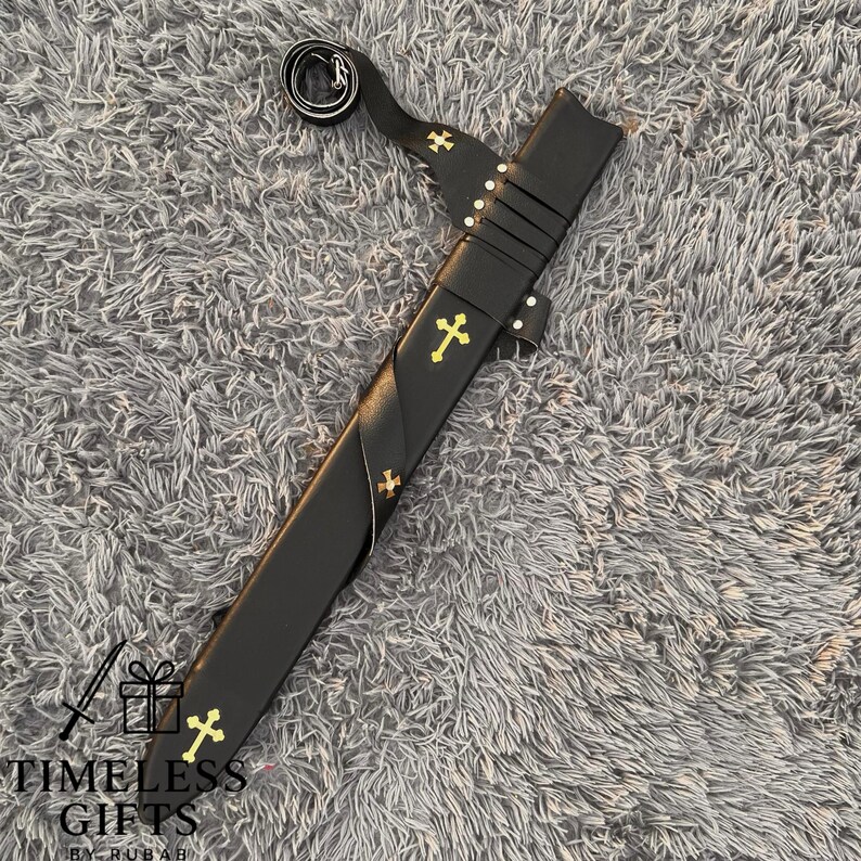 May include: A black sword with a leather sheath, featuring gold cross accents. The sword has a curved handle and a strap. The sheath is adorned with gold crosses. The text "TIMELESS GIFTS BY RUBAH" is visible at the bottom.