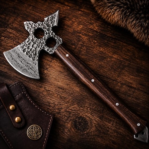 May include: A handcrafted axe with a dark wooden handle and a textured, patterned steel blade. The axe head features decorative cutouts. The handle is adorned with small, white decorative pins. The axe is displayed on a dark wood surface.
