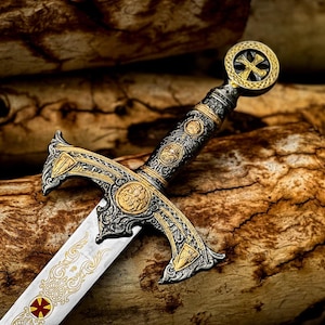 May include: An ornate sword with a silver blade and gold detailing. The hilt features intricate designs and a cross-shaped pommel. The sword rests on a wooden surface, highlighting its detailed craftsmanship.