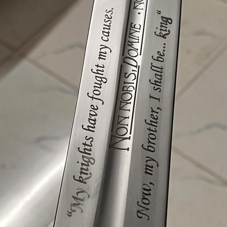 May include: Close-up of a silver sword blade with engraved text. The blade features the phrases "My knights have fought my causes," "NON NOBIS, DOMINE," and "Now, my brother, I shall be... king." The text is in a dark color, contrasting with the shiny metal.