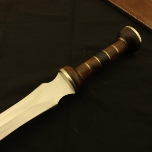 May include: A close-up of a Roman gladius sword. The blade is silver, and the handle is made of dark wood with brass accents. A brown leather sheath is visible in the background.