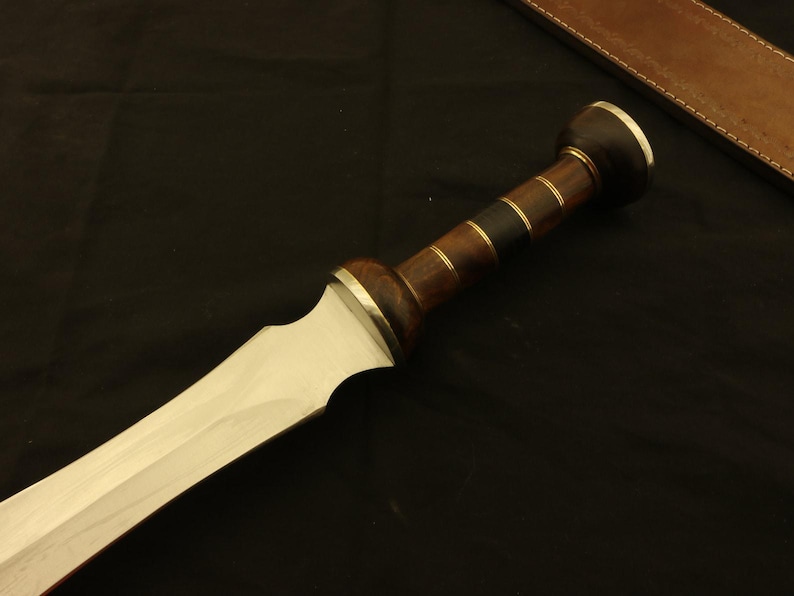 May include: A close-up of a Roman gladius sword. The blade is silver, and the handle is made of dark wood with brass accents. A brown leather sheath is visible in the background.