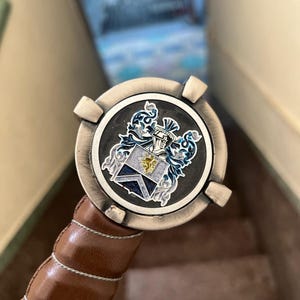 May include: A decorative sword hilt featuring a round, silver-toned pommel with a detailed crest design in blue, gold, and white. The handle is wrapped in brown leather with white stitching.