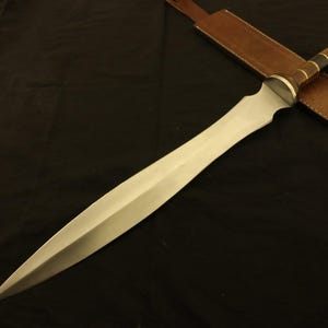 May include: A Gladius sword with a polished steel blade and a wooden handle with brass accents. The sword rests on a dark surface, with a brown leather sheath visible in the background. The blade is approximately 24 inches long.
