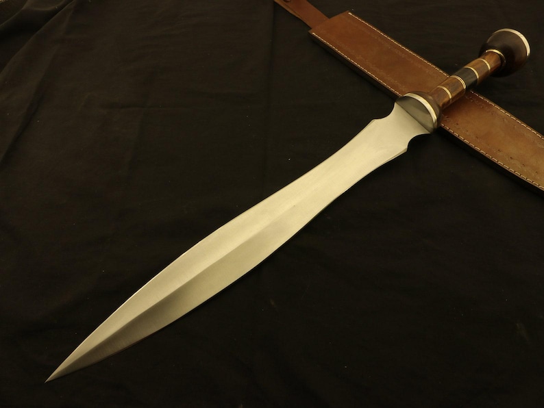 May include: A Gladius sword with a polished steel blade and a wooden handle with brass accents. The sword rests on a dark surface, with a brown leather sheath visible in the background. The blade is approximately 24 inches long.