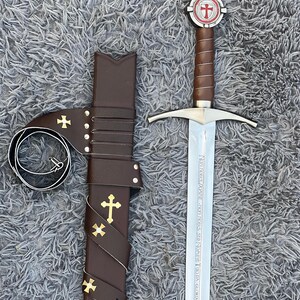 May include: A medieval sword with a brown leather-wrapped handle and a silver blade, next to a matching brown leather sheath. The sword's hilt features a red and white cross emblem. The sheath is adorned with gold crosses.