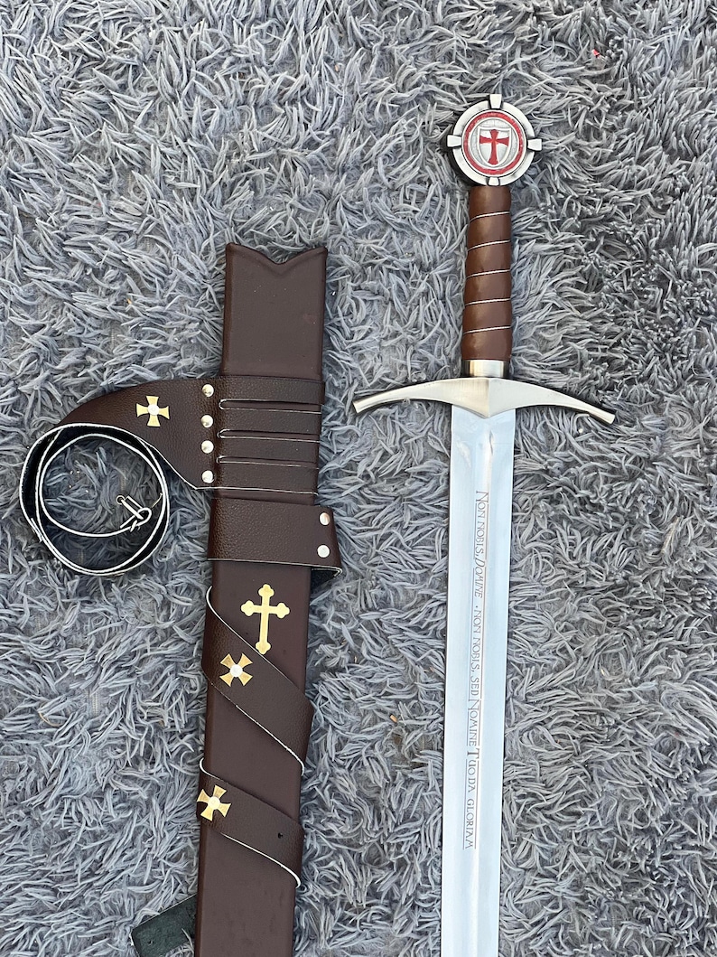 May include: A medieval sword with a brown leather-wrapped handle and a silver blade, next to a matching brown leather sheath. The sword's hilt features a red and white cross emblem. The sheath is adorned with gold crosses.