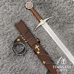 May include: A medieval-style sword with a brown leather-wrapped handle and a silver blade. The sword is displayed with its brown leather sheath, adorned with gold crosses. The sword's hilt features a decorative emblem. The image is set against a gray, textured background.