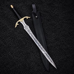 May include: A silver-toned sword featuring a gold-colored crossguard with ornate scrollwork and a black wrapped handle. The blade is decorated with an etched leaf vine pattern and is accompanied by a black leather scabbard with a metal rivet.