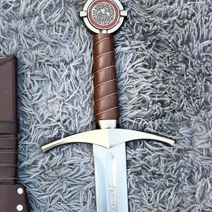 May include: A medieval-style sword with a silver blade and a brown leather-wrapped handle. The hilt features a circular emblem with red and white accents. The sword is displayed on a gray, textured surface.