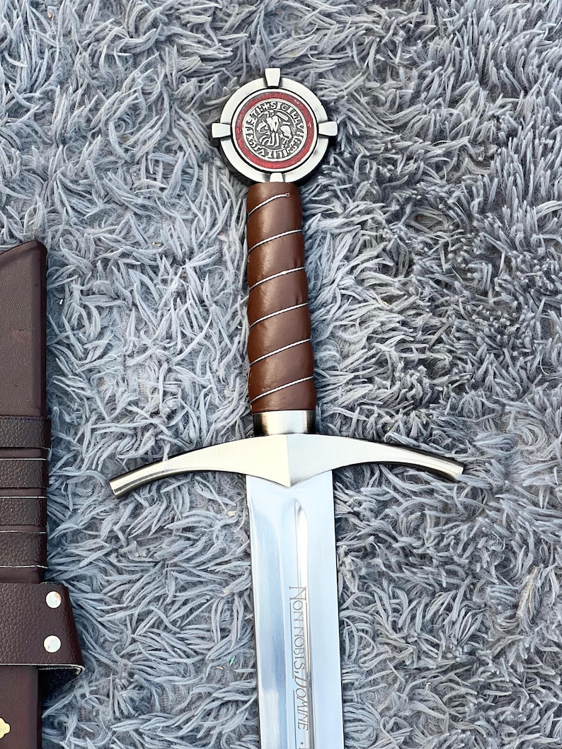May include: A medieval-style sword with a silver blade and a brown leather-wrapped handle. The hilt features a circular emblem with red and white accents. The sword is displayed on a gray, textured surface.