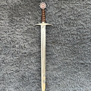 May include: A long, silver sword with a brown leather-wrapped handle and a circular pommel featuring a red cross design. The blade has a pointed tip and a crossguard. The sword is displayed on a gray, textured surface.