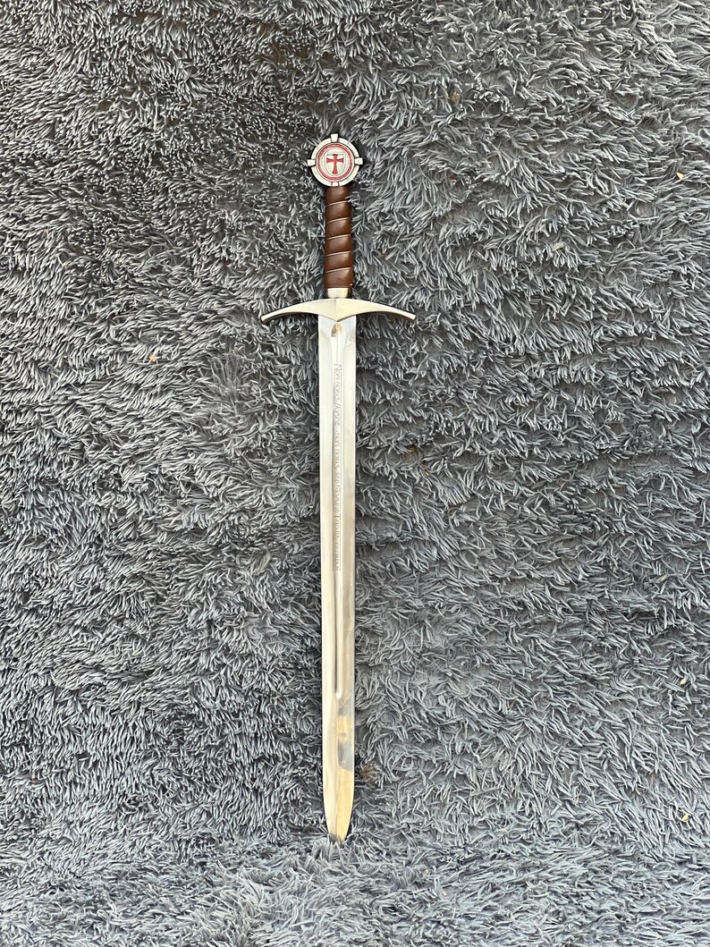 May include: A long, silver sword with a brown leather-wrapped handle and a circular pommel featuring a red cross design. The blade has a pointed tip and a crossguard. The sword is displayed on a gray, textured surface.