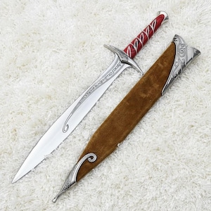 Puede incluir: Una espada plateada con empuñadura roja y plateada y una vaina marrón. La espada tiene una hoja larga y puntiaguda con detalles grabados. La vaina tiene detalles plateados con un diseño decorativo.
