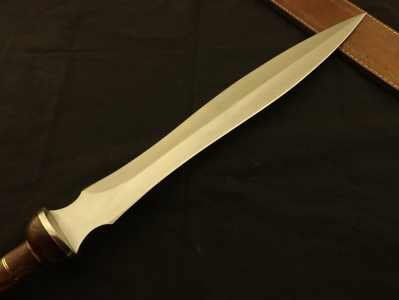 May include: A long, silver-colored blade with a wooden handle and a brass accent. The blade has a sharp point and a wide, flat surface. The handle is dark brown. The sword is approximately 24 inches long.