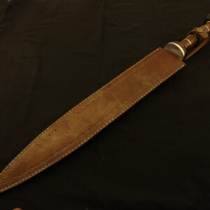 May include: A brown leather sheath houses a dagger with a dark handle and silver accents. The dagger's handle has a rounded end with a dark brown cap. The sheath is stitched with white thread.