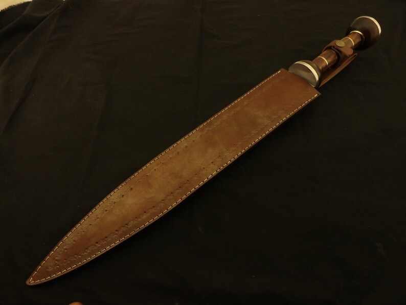 May include: A brown leather sheath houses a dagger with a dark handle and silver accents. The dagger's handle has a rounded end with a dark brown cap. The sheath is stitched with white thread.