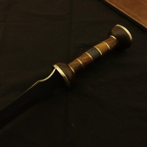 May include: A close-up of a gladius sword. The sword features a dark blade and a wooden handle with brass accents. The handle has a cylindrical shape with a rounded pommel. A leather sheath is visible in the background.