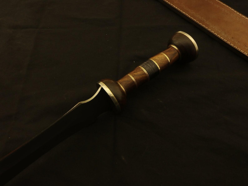 May include: A close-up of a gladius sword. The sword features a dark blade and a wooden handle with brass accents. The handle has a cylindrical shape with a rounded pommel. A leather sheath is visible in the background.
