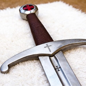 Op de afbeelding: Een stalen zwaard met een bruin omwikkeld handvat, een pareerstang met een gegraveerd kruis en een pommel met een rood kruisembleem. Het lemmet bevat een klein gegraveerd kruissymbool bij het gevest en heeft een metalen afwerking.