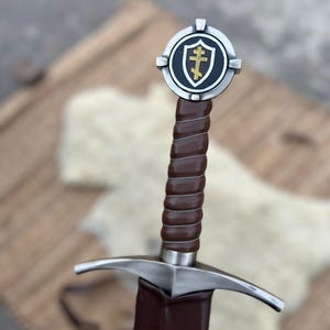 May include: A medieval sword with a brown leather-wrapped handle and a silver crossguard. The pommel features a shield emblem with a gold cross on a black background, framed by a silver circle. The sword is a historical replica.