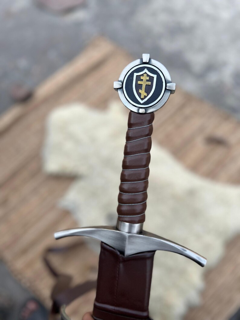 May include: A medieval sword with a brown leather-wrapped handle and a silver crossguard. The pommel features a shield emblem with a gold cross on a black background, framed by a silver circle. The sword is a historical replica.