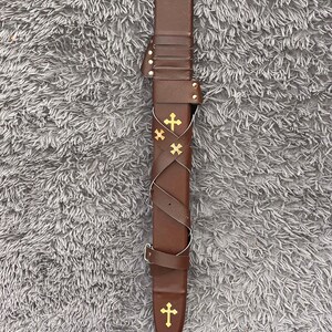 May include: A brown leather sword sheath with gold cross embellishments. The sheath has a decorative strap and buckle design. The sheath is designed to hold a sword. The sheath is on a gray surface.