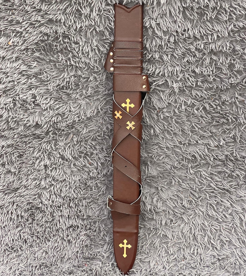 May include: A brown leather sword sheath with gold cross embellishments. The sheath has a decorative strap and buckle design. The sheath is designed to hold a sword. The sheath is on a gray surface.
